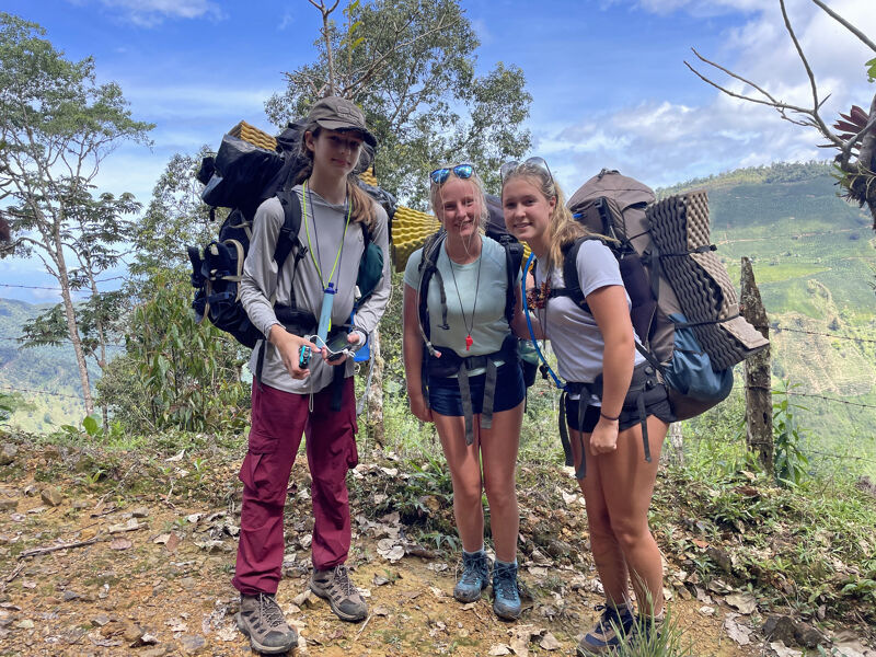 Three young women are standing outside, likely on a hiking trail, each carrying a large backpack. The woman on the left is wearing a hat and red pants. The woman in the center has blonde hair and is wearing shorts. The woman on the right has blonde hair in a ponytail and is also wearing shorts. They appear to be taking a break during their hike.
